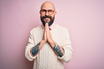 Handsome bald man with beard and tattoo wearing glasses and sweater over pink background praying with hands together asking for forgiveness smiling confident.