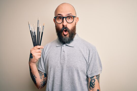 Handsome bald artist man with beard and tattoo painting using painter brushes scared in shock with a surprise face, afraid and excited with fear expression