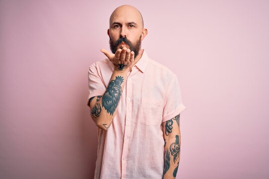 Handsome bald man with beard and tattoo wearing casual shirt over isolated pink background looking at the camera blowing a kiss with hand on air being lovely and sexy. Love expression.