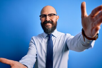 Handsome business bald man with beard wearing elegant tie and glasses over blue background looking at the camera smiling with open arms for hug. Cheerful expression embracing happiness.