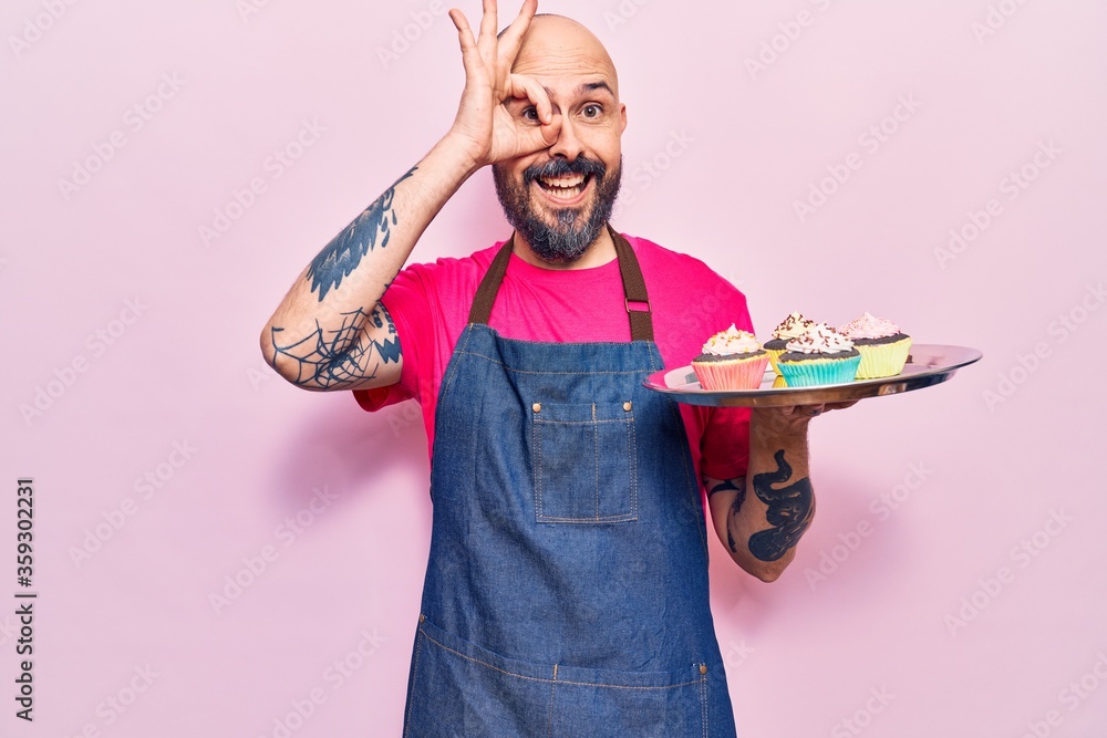 Poster Young handsome man wearing apron holding cupcake smiling happy doing ok sign with hand on eye looking through fingers