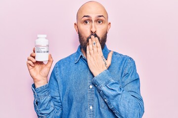 Young handsome man holding pills covering mouth with hand, shocked and afraid for mistake....