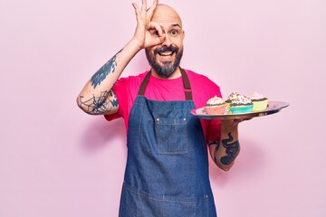 Young handsome man wearing apron holding cupcake smiling happy doing ok sign with hand on eye looking through fingers