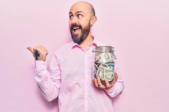 Young handsome man holding jar with savings pointing thumb up to the side smiling happy with open mouth