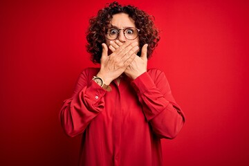 Middle age beautiful curly hair woman wearing casual shirt and glasses over red background shocked covering mouth with hands for mistake. Secret concept.