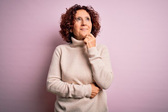 Middle age beautiful curly hair woman wearing casual turtleneck sweater over pink background with hand on chin thinking about question, pensive expression. Smiling and thoughtful face. Doubt concept.