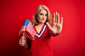 Middle age blonde patriotic woman holding united states flags celebrating independence day with open hand doing stop sign with serious and confident expression, defense gesture