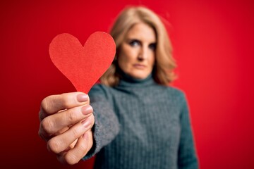 Middle age beautiful blonde romantic woman holding red paper heart celebrating valentine day with a confident expression on smart face thinking serious