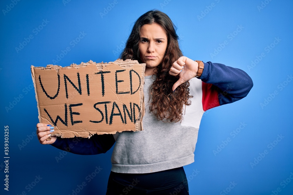 Sticker Beautiful activist woman holding banner with united stand message over blue background with angry face, negative sign showing dislike with thumbs down, rejection concept