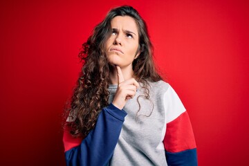 Young beautiful woman with curly hair wearing casual sweatshirt over isolated red background Thinking concentrated about doubt with finger on chin and looking up wondering