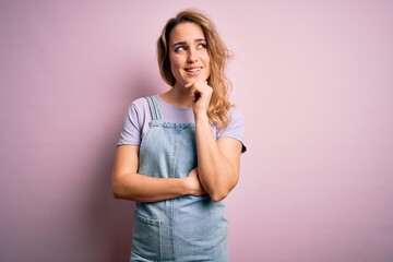 Young beautiful blonde woman wearing casual denim overalls standing over pink background with hand on chin thinking about question, pensive expression. Smiling with thoughtful face. Doubt concept.
