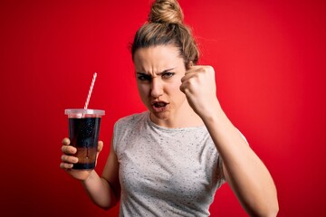 Beautiful blonde woman drinking cola fizzy beverage to refreshment over red background annoyed and frustrated shouting with anger, crazy and yelling with raised hand, anger concept