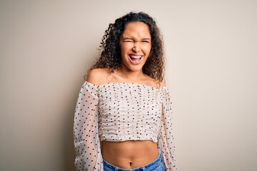Young beautiful woman with curly hair wearing casual t-shirt standing over white background winking...
