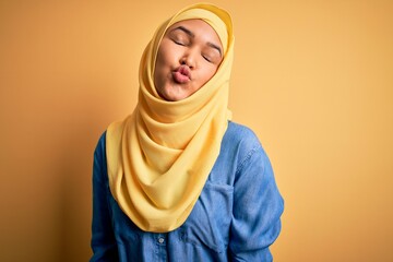 Young beautiful woman with curly hair wearing arab traditional hijab over yellow background looking at the camera blowing a kiss on air being lovely and sexy. Love expression.