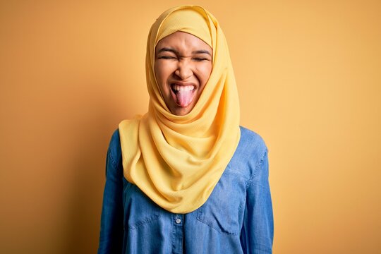 Young Beautiful Woman With Curly Hair Wearing Arab Traditional Hijab Over Yellow Background Sticking Tongue Out Happy With Funny Expression. Emotion Concept.