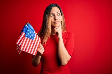 Young beautiful blonde patriotic woman with blue eyes holding united states flags serious face thinking about question, very confused idea