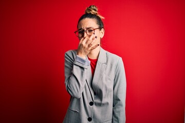 Young beautiful blonde businesswoman with blue eyes wearing glasses and jacket smelling something stinky and disgusting, intolerable smell, holding breath with fingers on nose. Bad smell