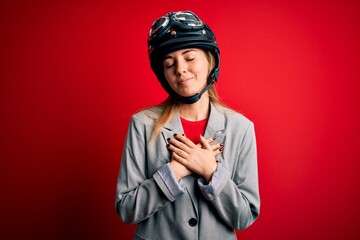 Young beautiful blonde motorcyclist woman wearing motorcycle helmet over red background smiling with hands on chest with closed eyes and grateful gesture on face. Health concept.