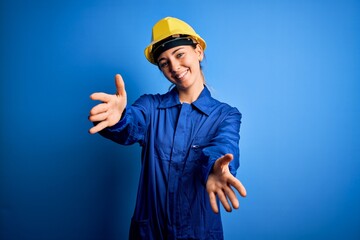 Young beautiful worker woman with blue eyes wearing security helmet and uniform looking at the camera smiling with open arms for hug. Cheerful expression embracing happiness.