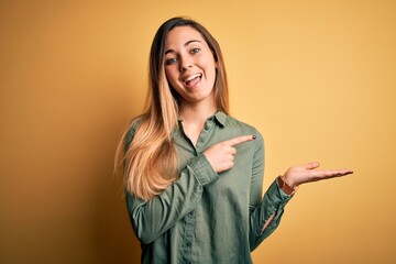 Young beautiful blonde woman with blue eyes wearing green shirt over yellow background amazed and smiling to the camera while presenting with hand and pointing with finger.