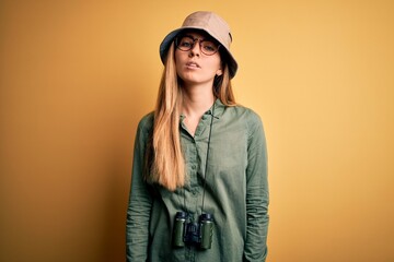 Beautiful blonde explorer woman with blue eyes wearing hat and glasses using binoculars Relaxed with serious expression on face. Simple and natural looking at the camera.