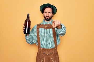 Young handsome man wearing tratidional german octoberfest custome and drinking beer from bottle pointing with finger to the camera and to you, hand sign, positive and confident gesture from the front