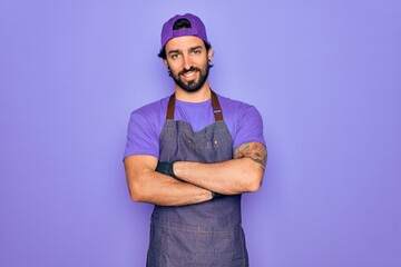 Young handsome hispanic man with bear wearing professional apron working as tattoo artist happy face smiling with crossed arms looking at the camera. Positive person.