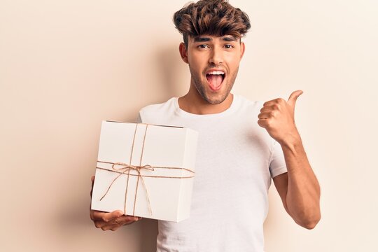 Young hispanic man holding gift pointing thumb up to the side smiling happy with open mouth