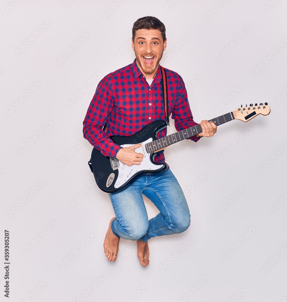 Wall mural Young handsome hispanic artist man playing electric guitar. Jumping with open mouth over isolated white background