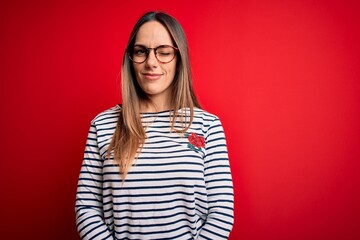 Young beautiful blonde woman with blue eyes wearing glasses standing over red background winking...