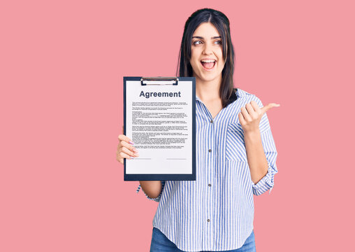 Young beautiful girl holding clipboard with agreement document pointing thumb up to the side smiling happy with open mouth