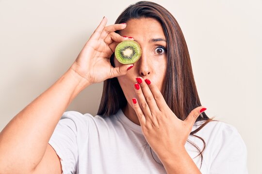 Young beautiful brunette woman holding kiwi covering mouth with hand, shocked and afraid for mistake. surprised expression
