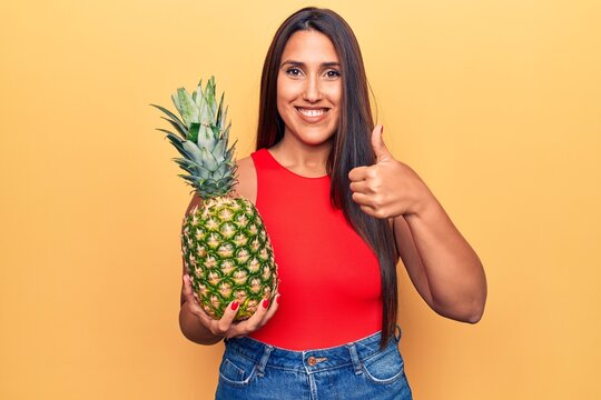 Young beautiful brunette woman holding pineapple smiling happy and positive, thumb up doing excellent and approval sign