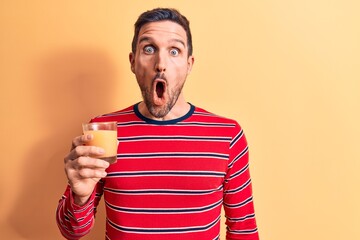 Young handsome man drinking glass of healthy orange juice over isolated yellow background scared...