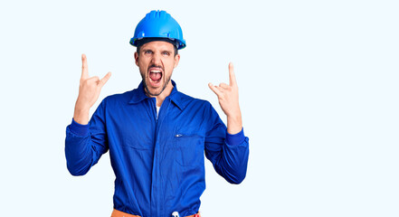 Young handsome man wearing worker uniform and hardhat shouting with crazy expression doing rock symbol with hands up. music star. heavy music concept.