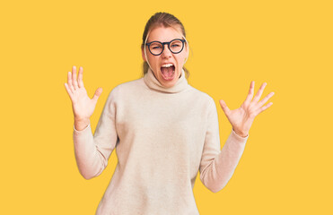Young beautiful blonde woman wearing turtleneck sweater and glasses crazy and mad shouting and...