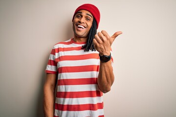 Fototapeta na wymiar Young handsome african american man with dreadlocks wearing striped t-shirt and wool hat smiling with happy face looking and pointing to the side with thumb up.