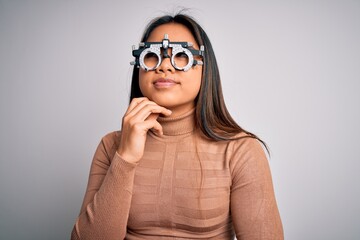 Young asian optical girl controlling eyesight using optometry glasses over white background with hand on chin thinking about question, pensive expression. Smiling with thoughtful face. Doubt concept.