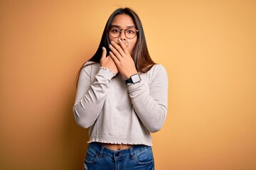 Young beautiful asian girl wearing casual sweater and glasses over yellow background shocked covering mouth with hands for mistake. Secret concept.