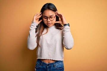 Young beautiful asian girl wearing casual sweater and glasses over yellow background with hand on head for pain in head because stress. Suffering migraine.