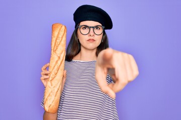 Young beautiful girl wearing fashion french beret holding fresh baked bread baguette pointing with finger to the camera and to you, hand sign, positive and confident gesture from the front