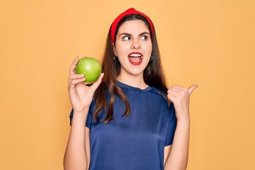 Young beautiful girl eating fresh organic healthy green apple over yellow background pointing and showing with thumb up to the side with happy face smiling