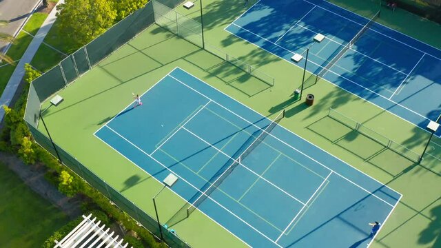 4K drone camera moves around a tennis court where there is an active game match. High quality 4k drone footage. Tennis court in the suburban area surrounded by the lavish green at sunset time.