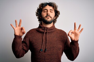 Young handsome man with beard wearing casual sweater standing over white background relax and smiling with eyes closed doing meditation gesture with fingers. Yoga concept.
