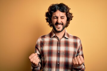 Young handsome man with beard wearing casual shirt standing over yellow background very happy and excited doing winner gesture with arms raised, smiling and screaming for success. Celebration concept.