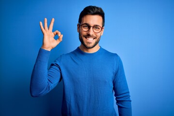 Young handsome man with beard wearing casual sweater and glasses over blue background smiling positive doing ok sign with hand and fingers. Successful expression. - obrazy, fototapety, plakaty