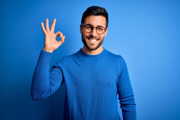 Young handsome man with beard wearing casual sweater and glasses over blue background smiling positive doing ok sign with hand and fingers. Successful expression.