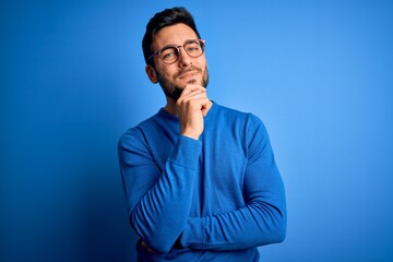 Young handsome man with beard wearing casual sweater and glasses over blue background looking...