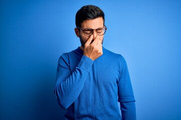 Obraz premium Young handsome man with beard wearing casual sweater and glasses over blue background tired rubbing nose and eyes feeling fatigue and headache. Stress and frustration concept.
