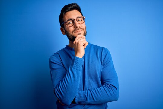 Young handsome man with beard wearing casual sweater and glasses over blue background looking confident at the camera smiling with crossed arms and hand raised on chin. Thinking positive.